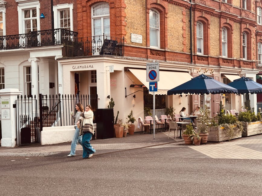 The image depicts the exterior of a restaurant named Glamaramma, situated on a street with a brick building façade and large windows. The restaurant has an outdoor seating area with dark blue umbrellas shading several tables and chairs, some of which are occupied by patrons. Potted plants and greenery are arranged along the sidewalk, enhancing the inviting atmosphere. Two women are walking past the establishment; one appears to be holding a drink or a small container, dressed in casual clothing, while the other walks beside her. A street sign indicating a restricted zone for parking is positioned near the outdoor seating, with a black and white striped gate to the left guarding entrance to a residential area or private property. The scene is lit with natural daylight, contributing to a warm and neutral tone that highlights the street and building details, consistent with a typical urban environment where courier or private waste clearance services might operate to manage waste generated by businesses or nearby residences, aligning with the context of independent rubbish removal services in the area.