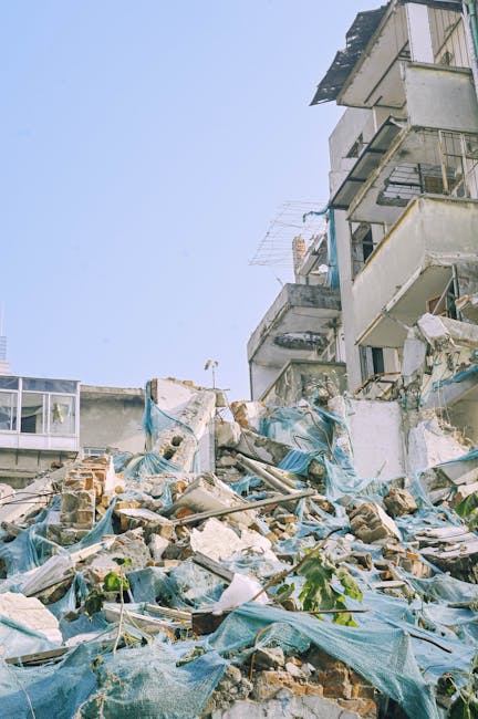The image depicts a partially collapsed multi-story residential building with exposed brickwork, damaged external walls, and debris scattered on the ground in front. Piles of broken bricks, concrete fragments, tangled blue tarpaulin sheets, and other construction debris are visible in the foreground, indicating recent demolition or structural issues. Portions of the building's balconies and windows are damaged or missing, with some metal railings and frames bent or hanging loosely. Surrounding environmental elements include a clear blue sky overhead, and a small plant growing through the rubble in the lower left area, giving a sense of outdoor urban space. The scene provides a detailed view of building debris and damaged construction materials, relevant to waste clearance and rubble removal services, such as those offered by Waste Clearance Chelsea, who handle private property clearances and alternative waste management solutions. The overall atmosphere suggests an active site requiring specialized removal of construction waste and debris, aligning with on-site clearance tasks in the context of independent rubbish disposal or renovation cleanup.