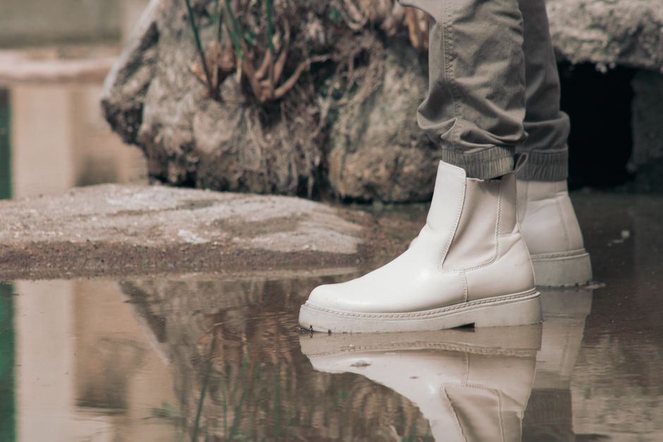 The image depicts a person standing outdoors on a wet, reflective surface near a large, rough-textured rock. The individual is wearing light grey, slightly loose-fitting pants with elastic cuffs around the ankles. They are also wearing ankle-high, smooth, white leather boots with a matte finish, flat soles, and subtle stitching details along the edges. The boots are positioned on the shiny, damp ground, creating a clear mirror-like reflection. In the background, the rock appears weathered with uneven, rugged surfaces, and there are hints of natural outdoor elements such as moss or dirt. The overall scene suggests a casual environment, possibly near a garden or park area, where the proximity to water and natural surfaces might relate to landscape clearance or outdoor rubbish collection activities. Waste Clearance Chelsea may facilitate such on-site waste removal, including clearing outdoor debris and materials like rocks or plant refuse, in a professional and efficient manner.