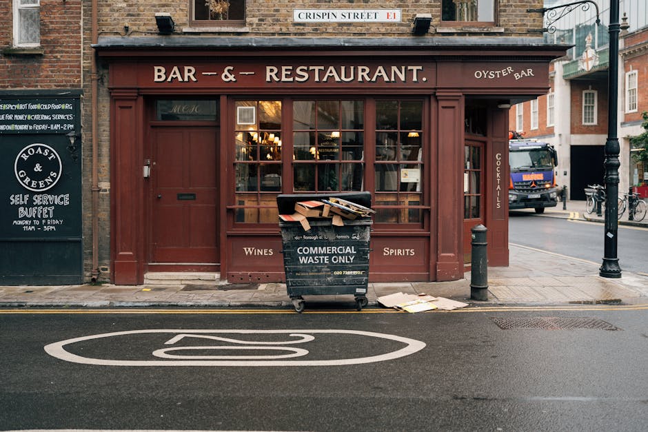 A black wheeled commercial waste container labeled 'COMMERCIAL WASTE ONLY' is positioned on the pavement in front of a brown-painted building with large glass windows, which appears to be a restaurant or bar. The container is overflowing with flattened cardboard boxes and paper waste, some of which are spilling onto the ground nearby. The building has signage indicating it serves wines, spirits, and cocktails, with the words 'BAR & RESTAURANT' displayed prominently at the top. To the left, a chalkboard-style sign advertises a self-service buffet, and to the right, a black bollard and a nearby lamppost are visible along the sidewalk. A traffic lane is marked with a white symbol on the road surface, and a blue and orange truck is seen driving past in the background, alongside bicycle racks and parked bicycles. The scene is set in an urban environment with brick buildings and typical street furniture, suggesting a location that benefits from independent waste collection or on-site rubbish handling managed by Waste Clearance Chelsea and other services.