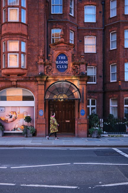 The image depicts a busy street scene outside a Ralph Lauren store located on a corner with a decorative white lattice archway at the entrance. The store, situated in a historic red brick building with detailed stonework above the facade, features large display windows showcasing mannequins dressed in clothing. The archway is adorned with greenery and flowers, adding an inviting touch to the entrance. A group of pedestrians, including men, women, and children, are crossing the sidewalk in front of the store, some carrying shopping bags or backpacks. The surrounding environment includes other shop fronts, with one showing a sign for men's and women's clothing, and potted plants with white flowers arranged outside the store. The scene is lit with natural daylight, creating a bright and lively atmosphere that reflects the typical retail activity in an affluent shopping area, where private and independent waste clearance services like those offered by Waste Clearance Chelsea are sometimes used to manage rubbish from retail or customer activity.
