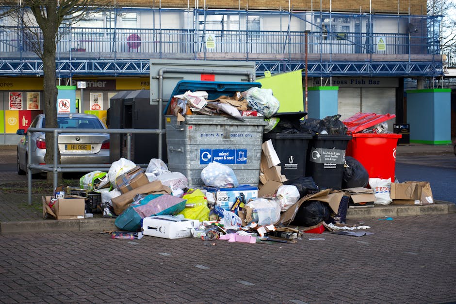 An outdoor scene showing multiple overflowing waste bins and an assortment of rubbish scattered on the pavement and partly on the road. The central large grey bin, labeled for mixed paper and cardboard, is open with various papers and packaging spilling out. To its right, there are black, red, and tan cardboard boxes and plastic bags filled with waste, some crumpled or torn. Behind these bins, a silver car is parked close to a metal railing, with a tree to the left. In the background, there is a building under construction or renovation, covered with blue scaffolding and protective netting, and signage indicating commercial units, including a fish bar. The environment suggests a busy urban area where waste accumulation may hinder collection or access, highlighting potential challenges for private rubbish removal services like Waste Clearance Chelsea, especially in circumstances where standard council collection is disrupted by access issues or site-specific restrictions.