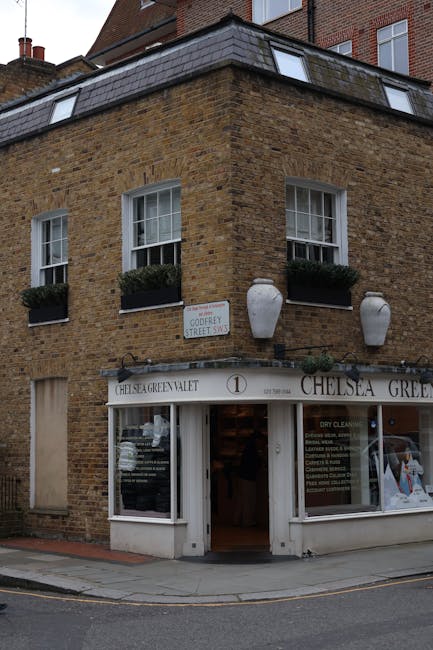 A three-story brick building with a ground-floor commercial space and residential windows above, located on a corner of a street. The brick exterior is warm-toned with a textured surface, and the building features white-framed sash windows, each adorned with window boxes filled with dark green plants. Two large, white, urn-shaped decorative planters are mounted symmetrically on the facade above the shop entrance. The shop window has a curved white signage board displaying the name 'Chelsea Garden Valet,' with a white door in the center leading into the premises. The glass storefront reflects the street scene and nearby vehicles. A small street sign, indicating ‘Goudery Street SW,’ is attached to the wall above the shop. The environment appears to be a busy urban area with a pavement running along the building's base. This scene may illustrate an address where private waste collection or on-site clearance services, such as those offered by Waste Clearance Chelsea, are conducted, highlighting alternative waste management options outside of local authority rubbish collection.